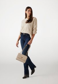 Strickpullover in Creme mit Blättermuster, dunkelblaue Schlagjeans und eine beigefarbene gesteppte Handtasche mit goldenen Kettenriemen. Dunkle Stiefel mit Absatz.