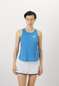Blå atletisk tanktopp med frontglidelås, med et strukturelt mønster og hvit logo. Kombinert med et hvitt skjørt med svart kant.