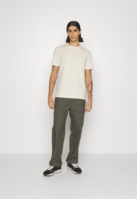 T-shirt beige à manches courtes associé à un pantalon vert olive, avec une coupe décontractée. Des baskets noires avec des lacets blancs complètent la tenue.