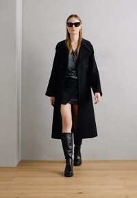Black oversized coat, gray satin top, black skirt, knee-high black boots. Model wears sunglasses, standing on light wood floor against a gray wall.