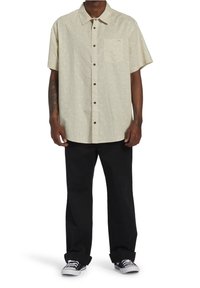 Korte mouwen, lichtbeige overhemd met knopen en een subtiel patroon, voorzak en bruine knopen, gecombineerd met zwarte broek en sneakers.