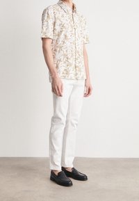 Camicia a maniche corte di un beige chiaro con motivo floreale, abbinata a pantaloni bianchi e mocassini neri. Tessuto morbido con vestibilità su misura.