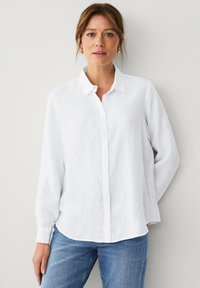 Camicia bianca in lino con bottoni, maniche lunghe, colletto classico e vestibilità rilassata, abbinata a jeans in denim blu.