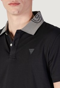 Polo negro hecho de tela suave, con un cuello gris de diseño intrincado, tapeta de tres botones y un logo triangular en el pecho.