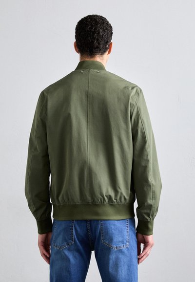 Zeleno-oljna bomber jakna z gladko teksturo, rebrastimi manšettami in robom ter minimalističnim dizajnom. Nošena čez modre kavbojke, videna s hrbta.
