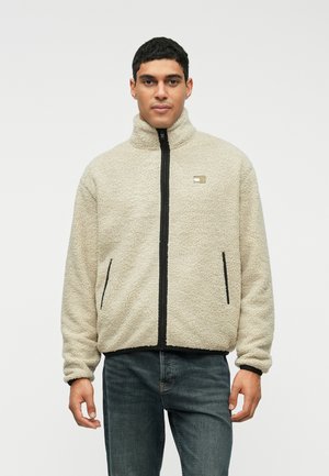 Homme portant une veste en polaire beige zippée avec un col haut et des poches bordées de noir, associée à un jean bleu foncé, se tenant contre un fond blanc.
