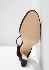 Chaussure noire à talon haut reposant sur le côté, montrant la semelle beige lisse et le talon texturé sur fond blanc.