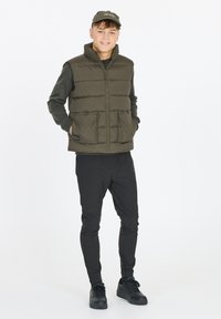 Olivgrüne Steppweste mit hohem Kragen, zwei vorderen Taschen, getragen über einem schwarzen Langarmshirt und kombiniert mit schwarzen Hosen und Sneakers.