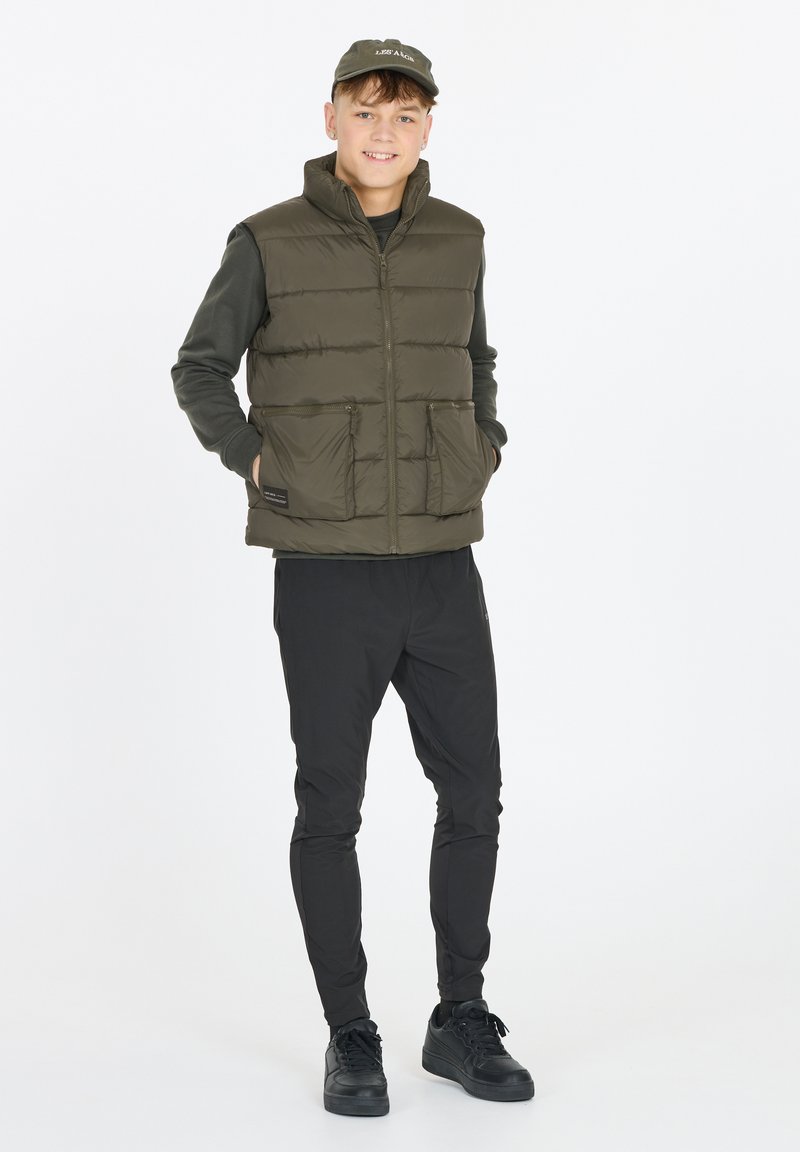 Olivgrüne Steppweste mit hohem Kragen, zwei vorderen Taschen, getragen über einem schwarzen Langarmshirt und kombiniert mit schwarzen Hosen und Sneakers.