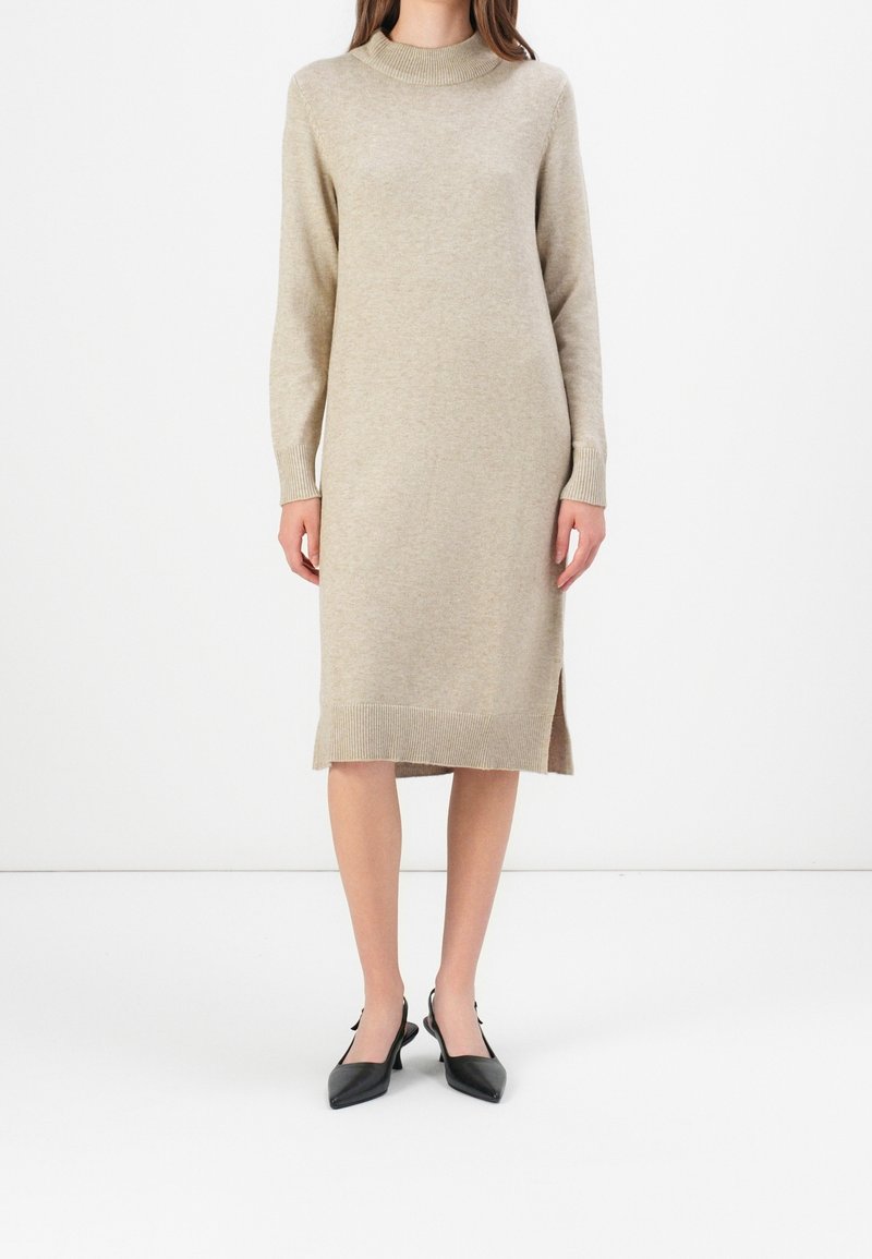 Femme portant une robe midi en maille beige à manches longues avec des fentes latérales et des escarpins noirs à bout pointu avec bride arrière, debout devant un fond blanc.