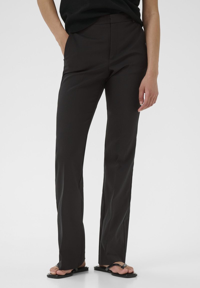 Pantalons noirs à jambe droite en tissu lisse, avec poches latérales et taille mi-haute, associés à des sandales plates.