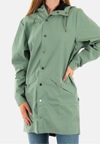 Vrouw met een knielange, saliegroene regenjas met drukknoopjes, klepzakken, capuchon, en met handen op de heupen en aan de zijkant.