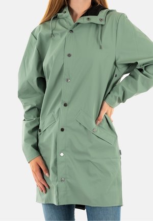 Femme portant un imperméable vert sauge arrive au genou avec des boutons-pression, des poches à rabat, une capuche, les mains sur les hanches et sur le côté.