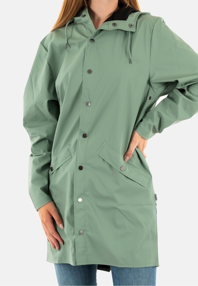 Vrouw met een knielange, saliegroene regenjas met drukknoopjes, klepzakken, capuchon, en met handen op de heupen en aan de zijkant.