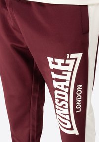 Bordo sportske hlače s bijelim bočnim akcentima i velikim logotipom "Lonsdale London", s glatkom teksturom i suženim dizajnom.