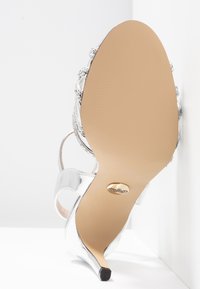 Sandale à talon haut argentée avec une semelle beige lisse et un petit emblème de marque près de la pointe du talon.