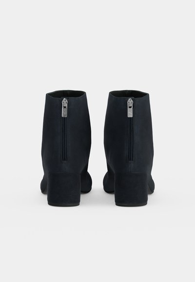 Paire de bottines noires à talons blocs, vues de dos, avec des fermetures éclair argentées centrales sur un fond clair uni.