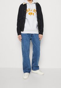 Person in einer schwarzen Reißverschlussjacke über einem hellgrauen Kapuzenpullover mit orangefarbenem Büffelmotiv, blauen Jeans und weißen Turnschuhen, steht an einer weißen Wand.