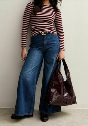 Žena v pruhovaném růžovo-černém topu s dlouhým rukávem, širokých modrých džínách, černých botách, držící velkou lesklou tmavě hnědou kabelku.