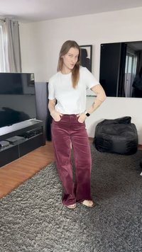 Weinrote Cordhose mit weitem Bein und strukturierter Oberfläche, kombiniert mit einem taillierten weißen T-Shirt. Die Kulisse zeigt einen modernen Wohnraum.