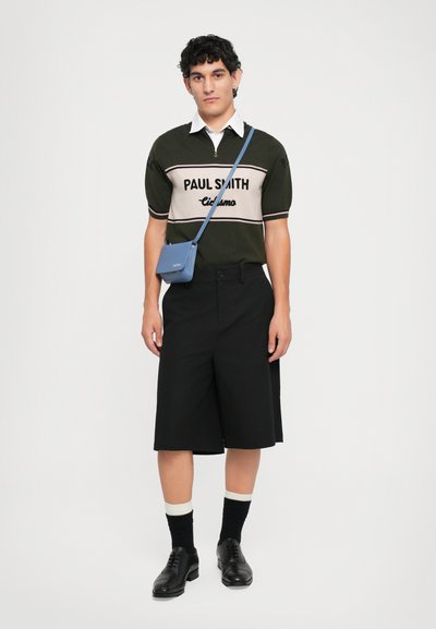 Junger Mann trägt einen Paul Smith Kurzarmpullover, schwarze lange Shorts, schwarze Socken mit weißen Streifen, schwarze Schuhe und eine blaue Umhängetasche.