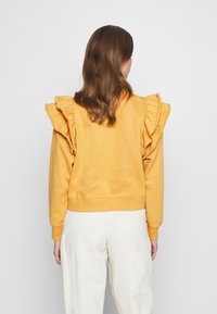 Žlutý svetr s výraznými volánkovými rukávy, žebrovanými manžetami a lemem. Pohled zezadu odhaluje hladkou texturu a uvolněný střih.