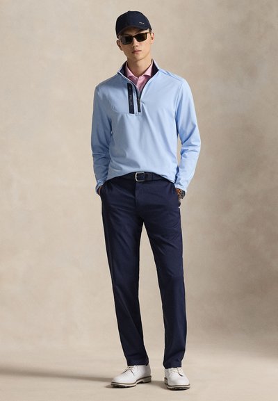 Ανοιχτό μπλε πουλόβερ με φερμουάρ στο γιακά, συνδυασμένο με σκούρο μπλε παντελόνι και λευκά παπούτσια, με αξεσουάρ γυαλιά ηλίου και σκούρο καπέλο.