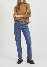 Bruine coltrui, blauwe rechte jeans, zwarte schoenen met gouden accenten. Zachte stof met een ontspannen pasvorm.