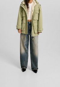 Olivově zelená parka s kapucí a Sherpa podšívkou, spojená se světle modrými širokými džínami. Černé boty s špičatou špicí dotvářejí outfit.