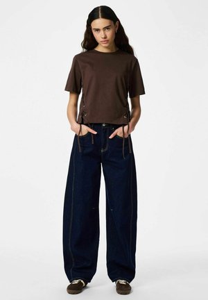 Ung kvinde med langt mørkt hår iført en brun kortærmet top med snøre detaljer i siden, vidde mørke jeans og brune sneakers, stående med hænderne i lommerne.