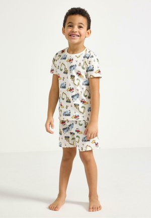 Smilende ung dreng står barfodet iført matchende hvid t-shirt og shorts med farverige byggepladskøretøjsmotiver.