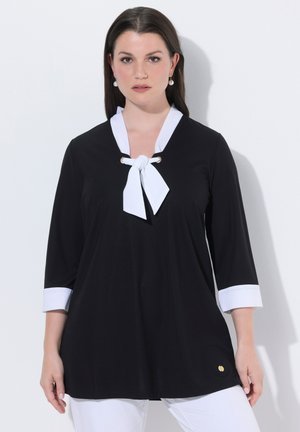 Kobieta ubrana w czarną bluzkę z długim rękawem, białym kołnierzykiem, krawatem i mankietami, w białych spodniach, stojąca na tle jednolitego białego tła.