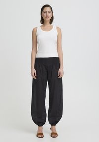 Kvinde iført en hvid ærmeløs top, løse sorte bukser med manchetter og brune sandaler med åbne tæer, stående mod en ensartet hvid baggrund.