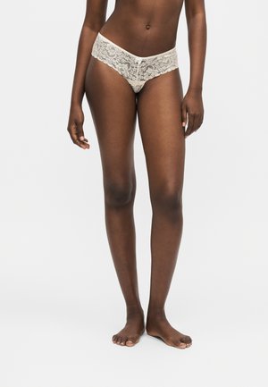 Mujer de pie descalza, con ropa interior blanca de encaje estilo hipster sobre un fondo claro y liso.