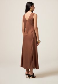 Robe sans manches marron avec un dos en V, faite d'un tissu texturé léger, silhouette fluide, portée avec des sandales à talons noirs.