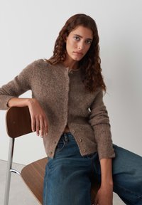 Bruine, fuzzy cardigan met een ronde halslijn, lange mouwen en knoopsluiting, gecombineerd met blauwe denim jeans. Houten stoel zichtbaar.
