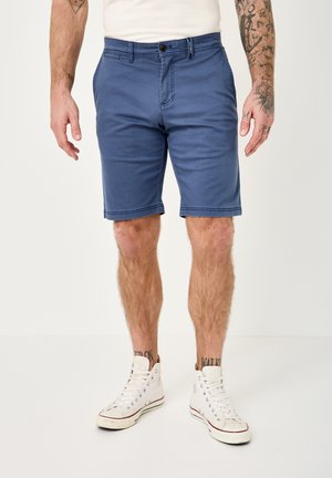 Man wearing blue knee-length shorts, white high-top sneakers, white t-shirt, with tattooed arms and legs visible from mid-thigh down.