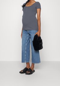 Mujer embarazada con una camiseta ajustada de manga corta gris, jeans anchos de mezclilla azul, sandalias negras de tipo deslizante y sosteniendo un bolso de pelo negro.