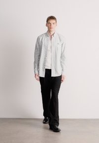 Πουκάμισο ανοιχτό με ανοιχτά μπλε ριγέ κουμπιά, φορεμένο πάνω από λευκό T-shirt, συνδυασμένο με μαύρο παντελόνι και μαύρα παπούτσια σε ένα μινιμαλιστικό περιβάλλον.