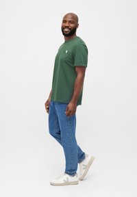 Groen t-shirt met korte mouwen en een borstzak, gecombineerd met lichtblauwe spijkerbroek en witte sneakers met beige accenten.