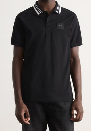 Homme portant un polo noir à manches courtes avec un col rayé blanc et un petit écusson logo "BOSS" sur le côté gauche de la poitrine.