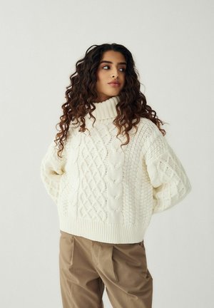 Moteris su ilgais garbanotais plaukais, dėvinti baltą pynuotą golfo megztinį ir smėlio spalvos kelnes, žiūrinti į šoną, rankos sukryžiuotos už nugaros.