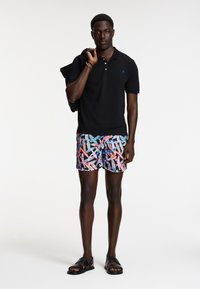 Čierne polo tričko, farebné plavky s vzorom a čierne sandále. Plavky sú zdobené mixom geometrických tvarov v ružovej, modrej a bielej farbe.