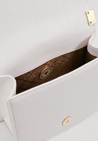 Vit läderhandväska med guldfärgad lås, knappstängning och brun insida med logomönster. Insidan har en slät textur.