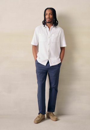 Man with shoulder-length dreadlocks wearing a white short-sleeve shirt, navy pants, and beige shoes, standing with hands in pockets.