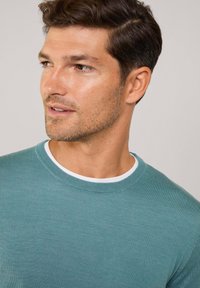 Turquoise crew-neck shirt made of knit fabric. Features a white collar accent and a textured surface. Model displays slight smile, facing right.