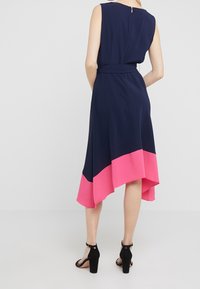 Marinblå ärmlös klänning med en rosa fåll, asymmetrisk silhuett, tygbälte i midjan och dragkedjestängning i ryggen. Svarta klackar.