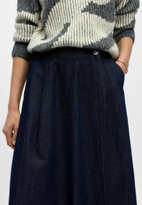 Kvindelig iført en mønstret grå og hvid sweater stoppet ned i en højtaljet mørkeblå denimnederdel med hænderne i lommerne.