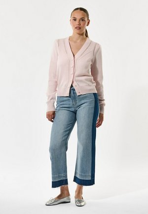 Donna in piedi che indossa un cardigan rosa pallido con bottoni, jeans cropped bicolore e ballerine argento su uno sfondo bianco semplice.
