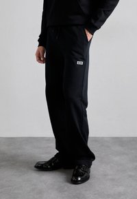 Czarne spodnie dresowe ze ściągaczem w talii i boczną kieszenią, z małą białą metką na górnej części uda. Zestawione z błyszczącymi czarnymi butami.